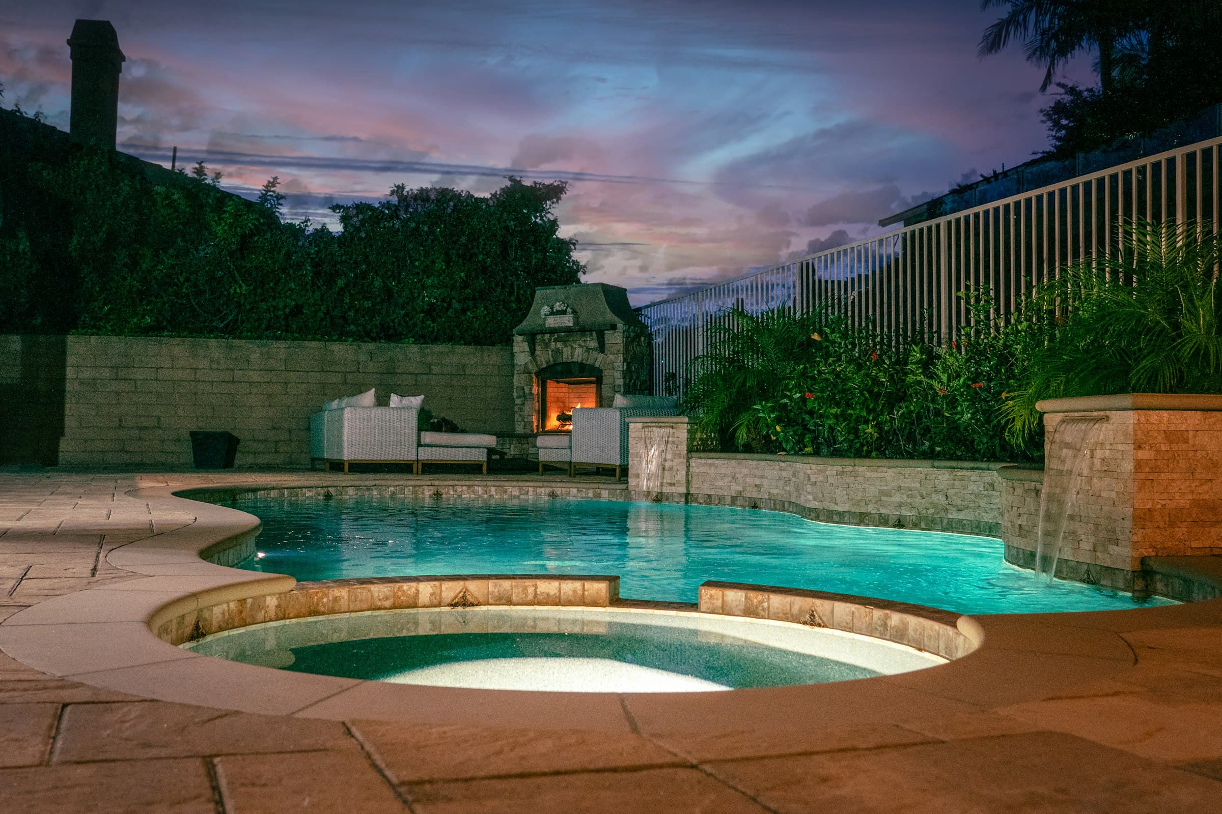 A backyard pool and spa lit at dusk, an outdoor fireplace behind them