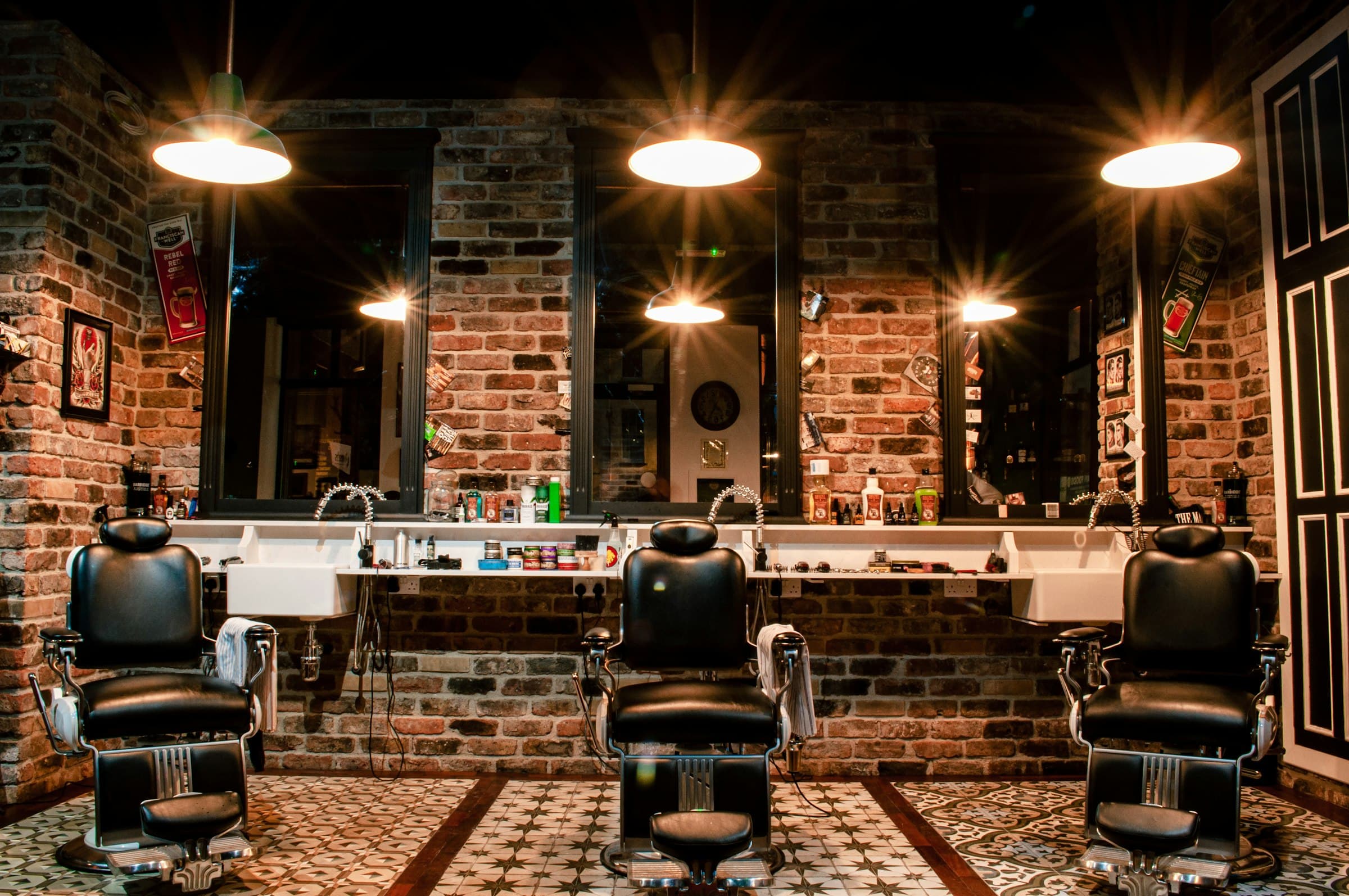 Three barber chairs before mirrors and a brick wall, under glowing lamps