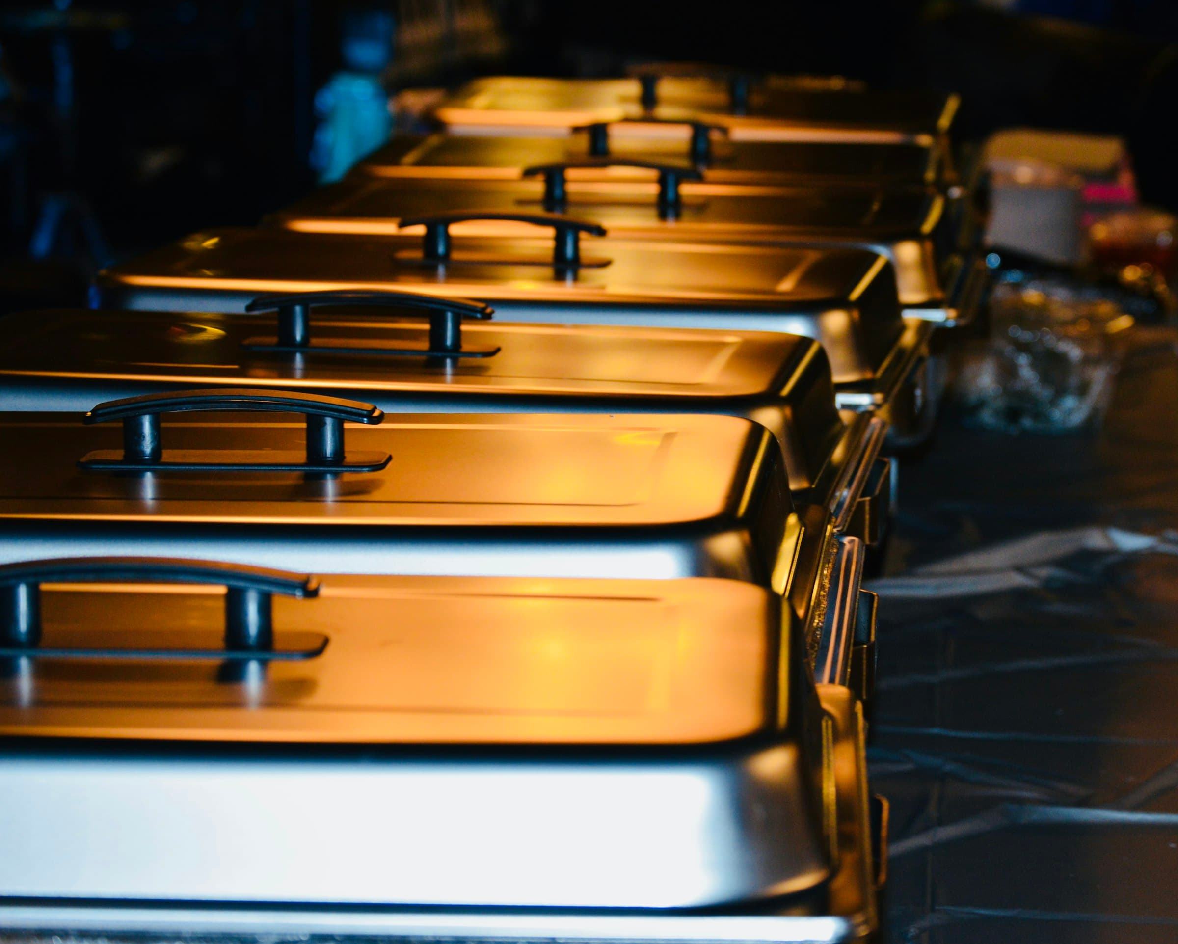 A row of polished chafing dishes on a buffet line, lit warm in the dark