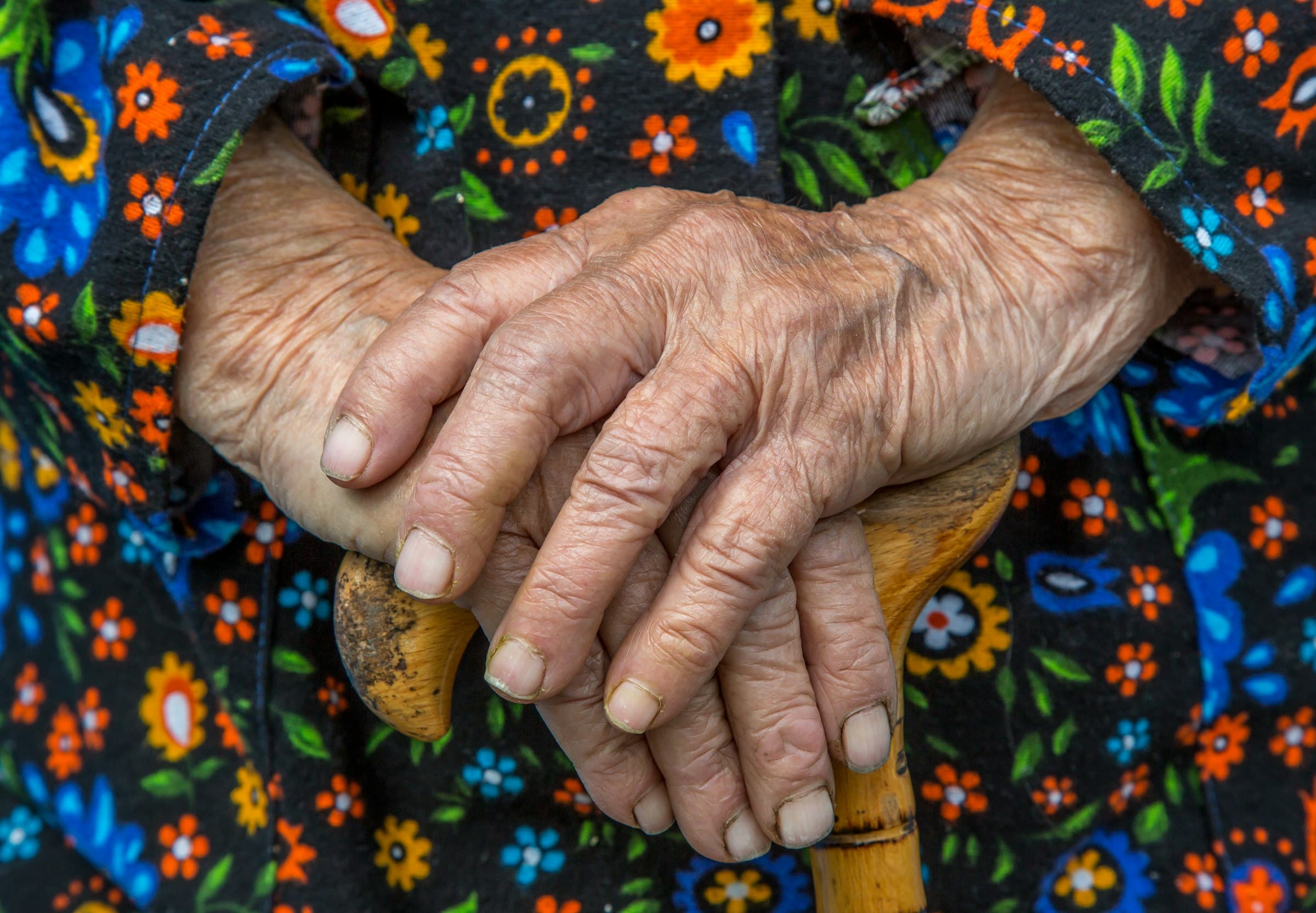 An older person's hands folded over the handle of a wooden cane, against a floral dress