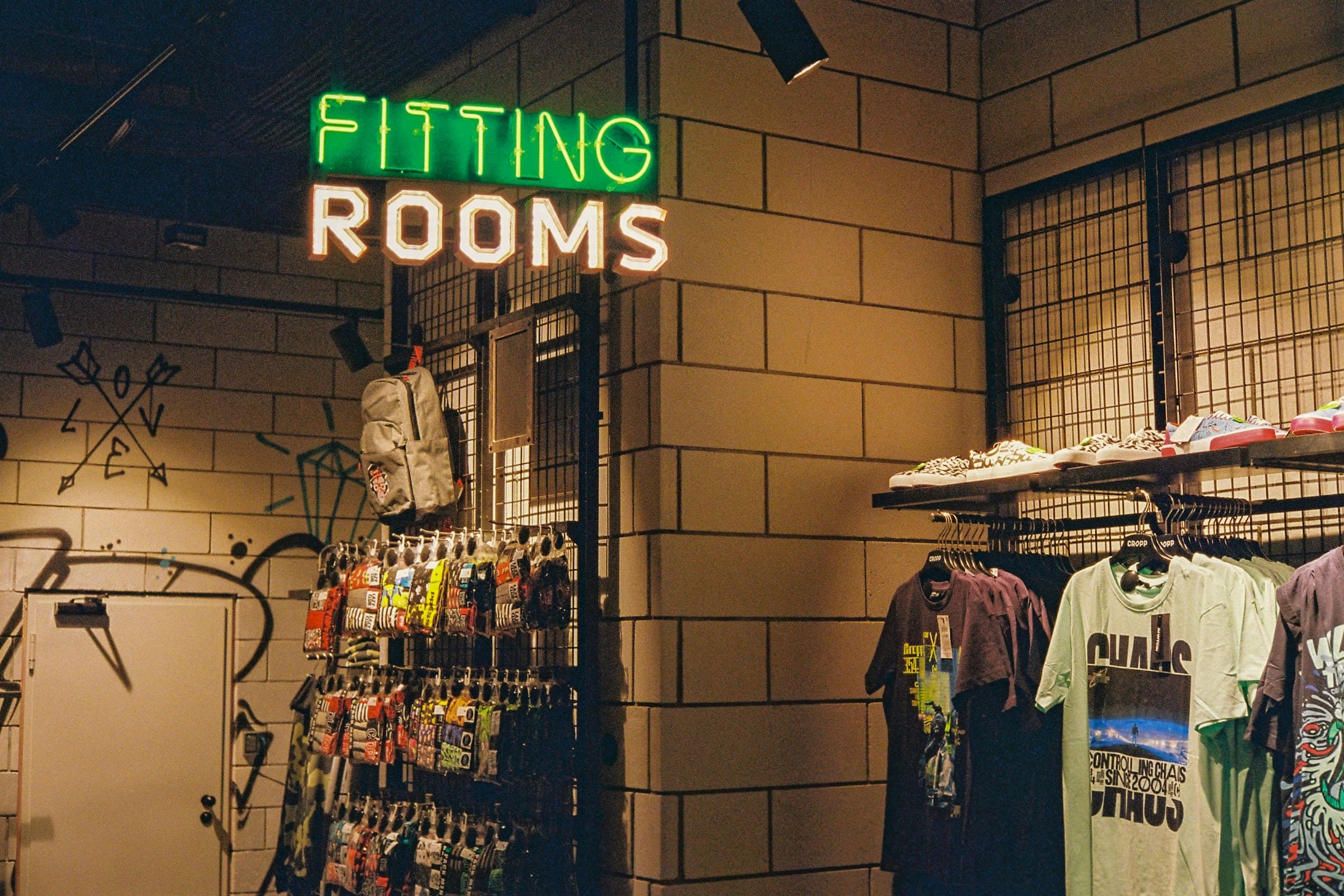 A neon FITTING ROOMS sign over racks of T-shirts in a clothing store