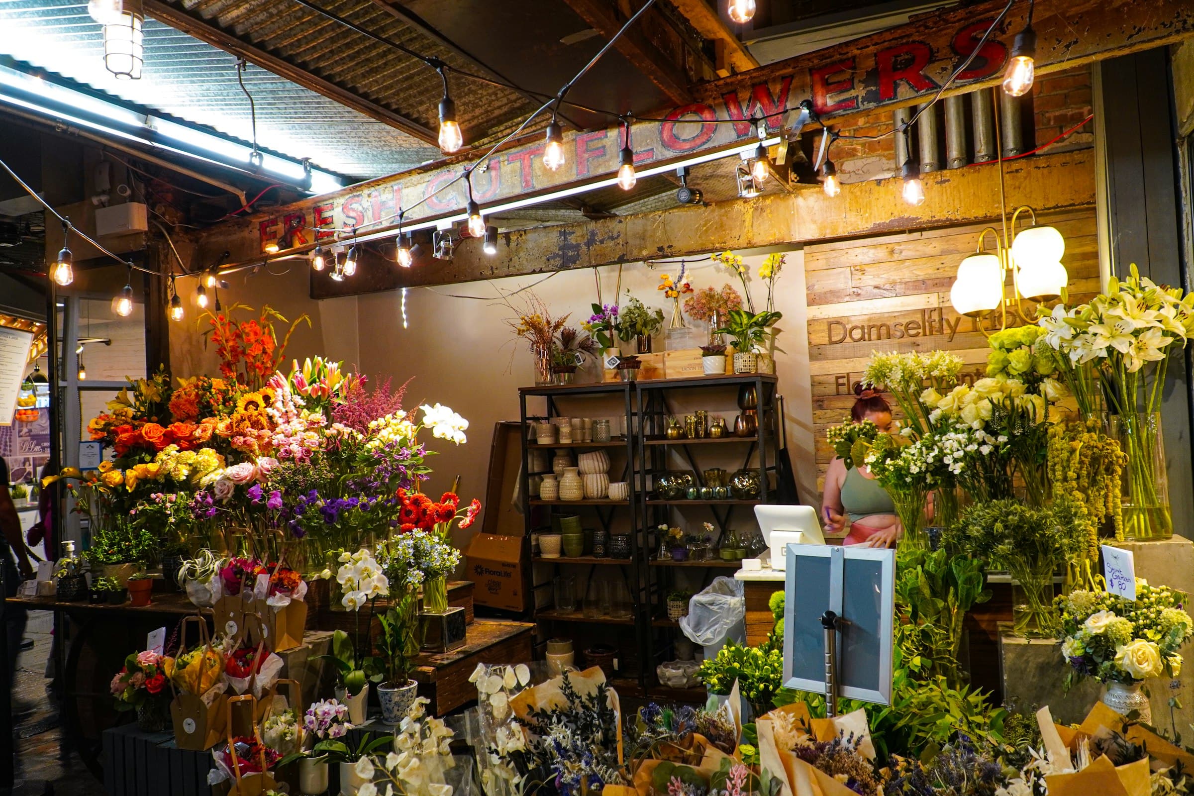 A flower shop crowded with buckets of flowers under strings of bulbs