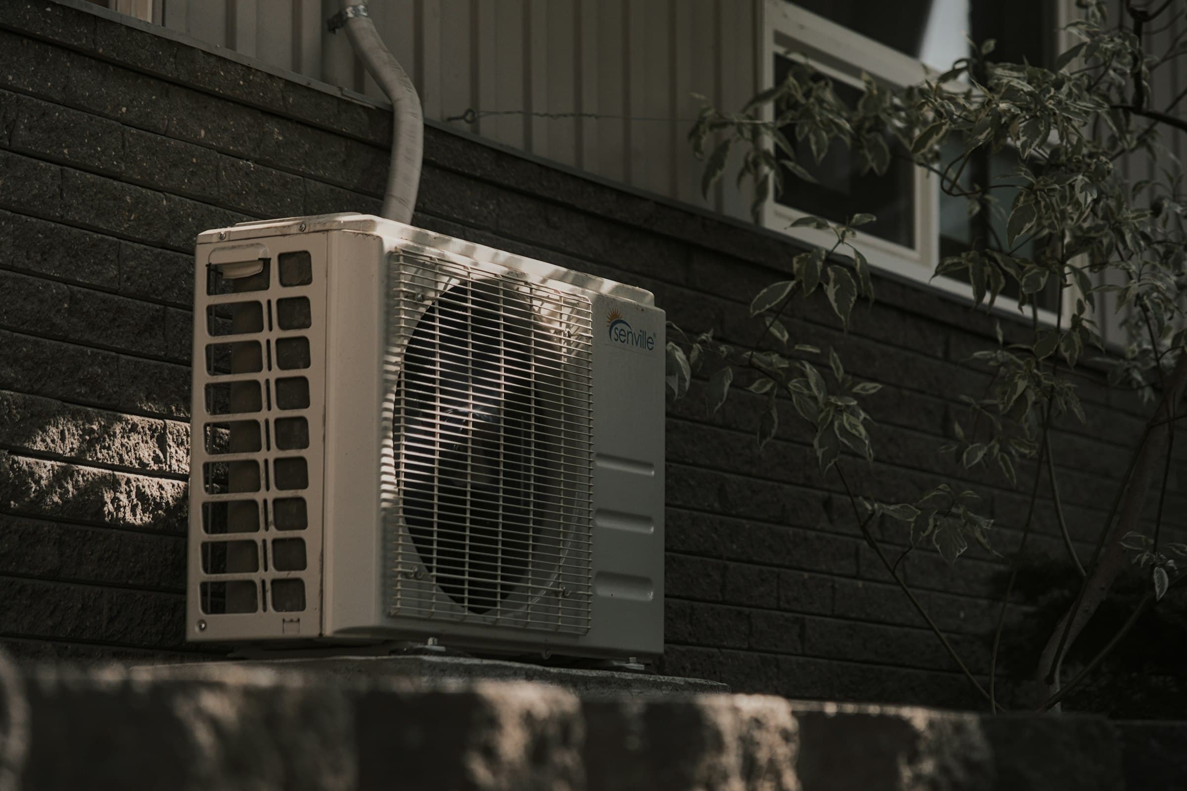An outdoor air conditioning unit against the dark brick wall of a house, a window and leaves above it