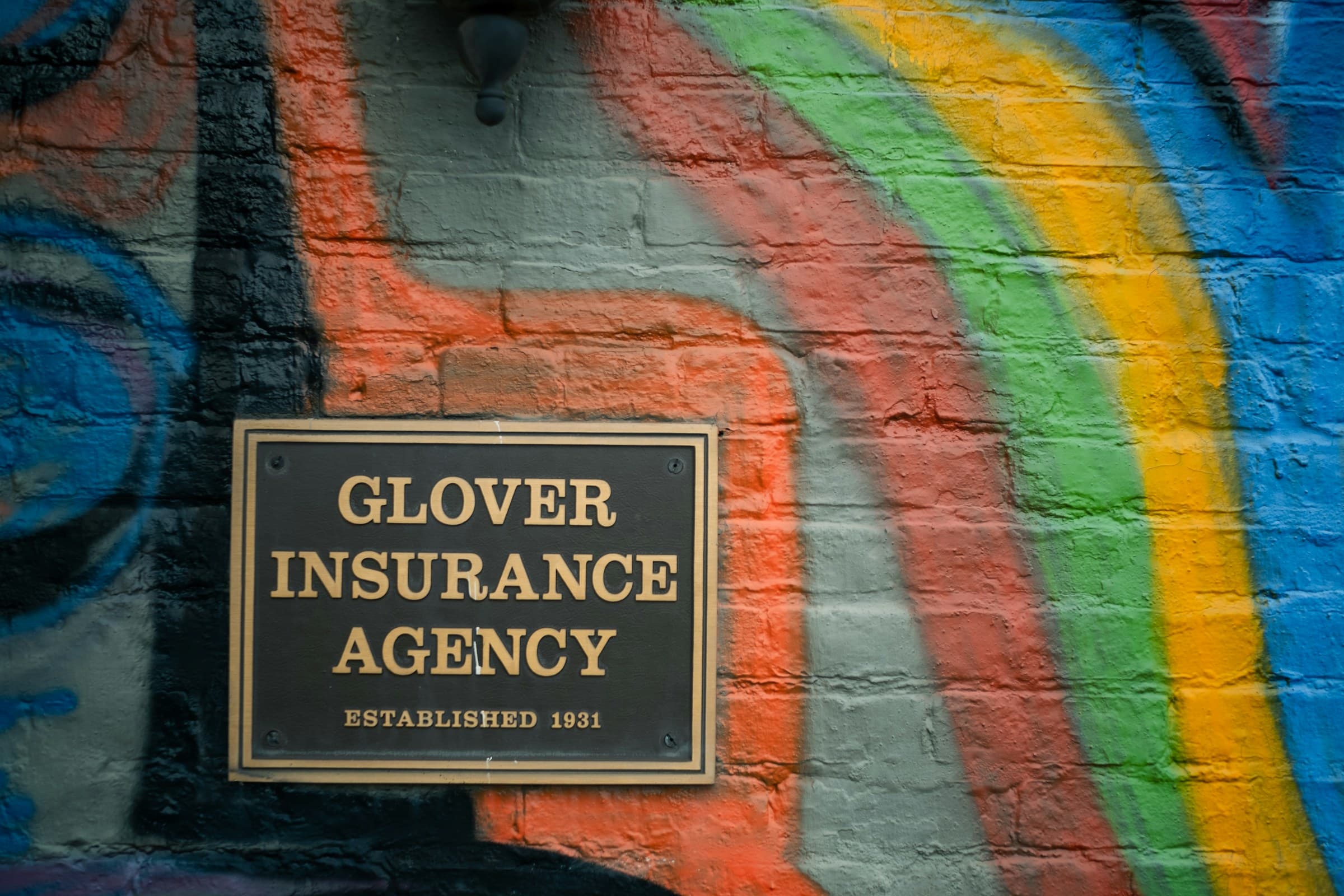A bronze plaque reading Glover Insurance Agency, Established 1931, on a brick wall painted with a rainbow