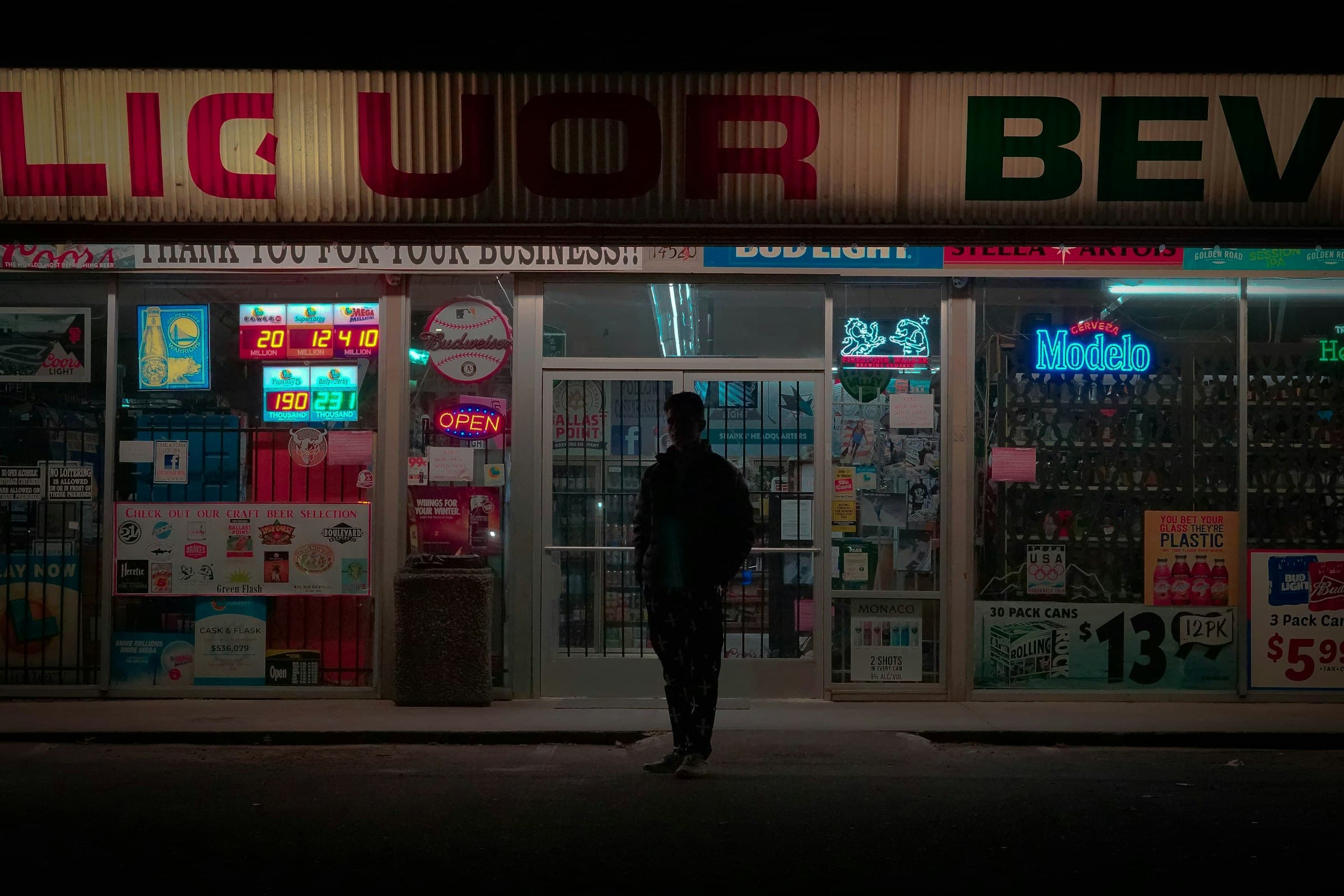A liquor store's lit windows at night, full of neon beer signs, a customer at the door