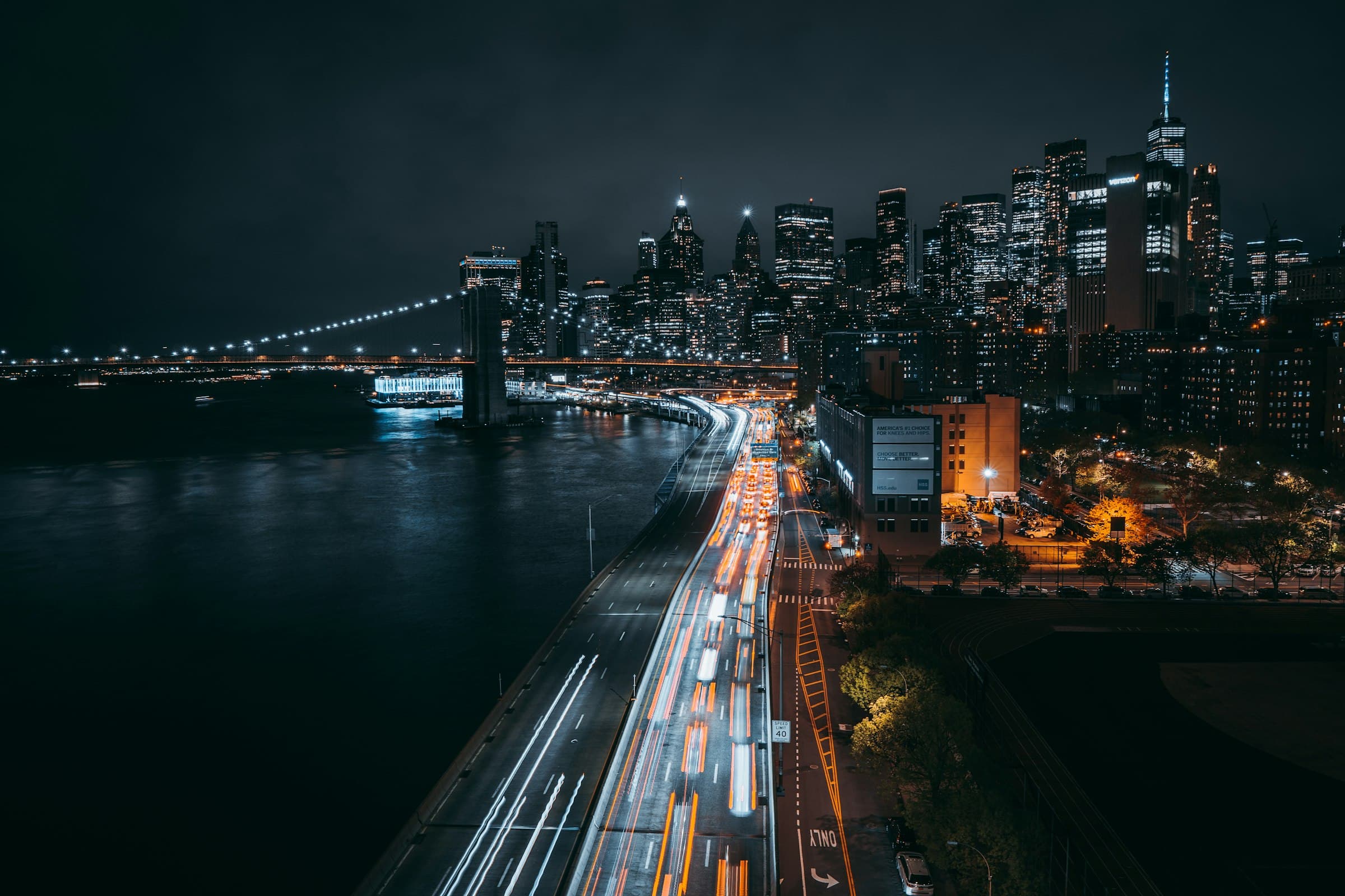 Lower Manhattan's towers at night from above, with the Brooklyn Bridge and streaks of traffic along the river