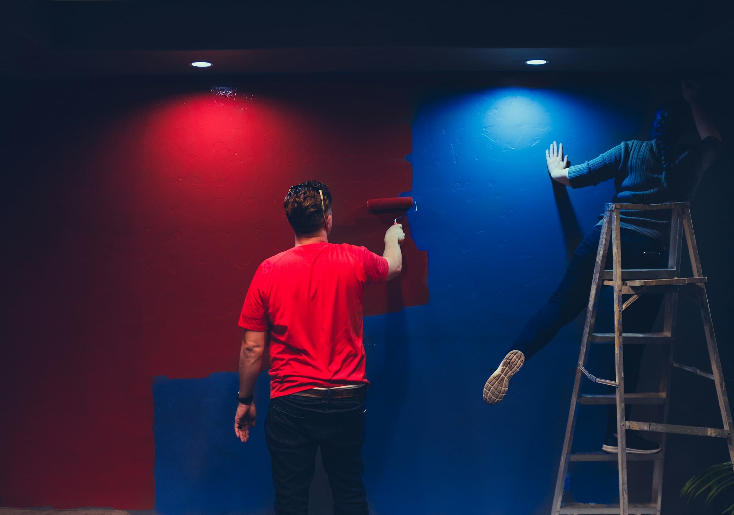 A painter rolling red paint over a blue wall under colored spotlights, a second painter up a ladder