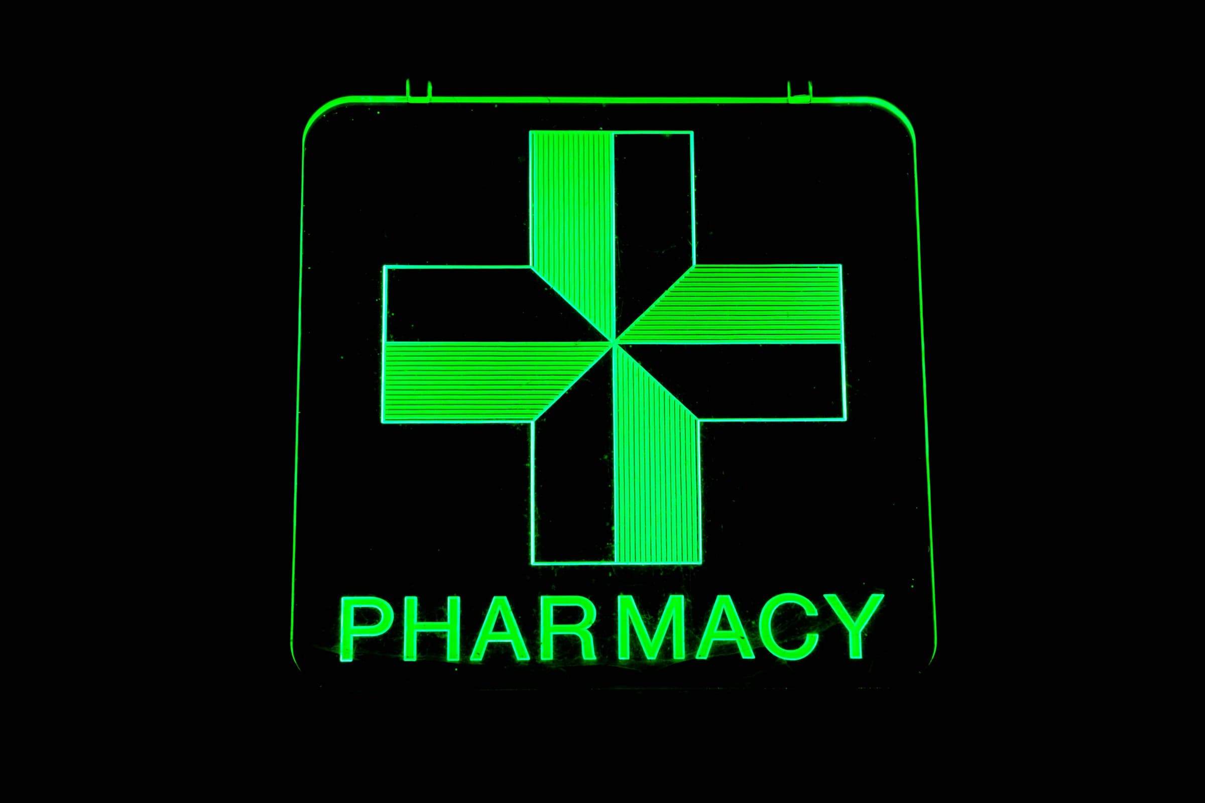 A green neon pharmacy cross above the word PHARMACY, glowing in the dark