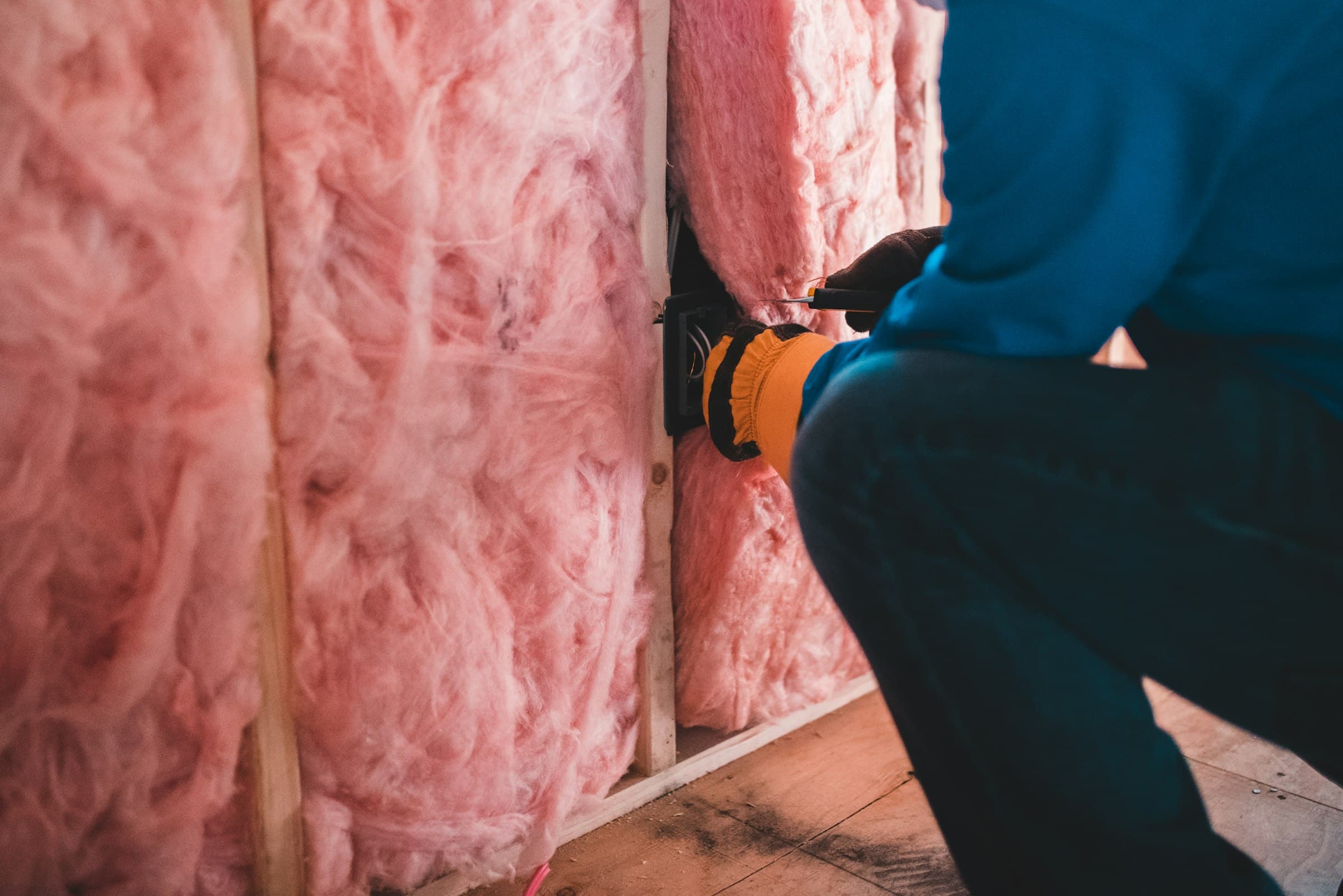 A worker fitting pink fiberglass insulation between wall studs