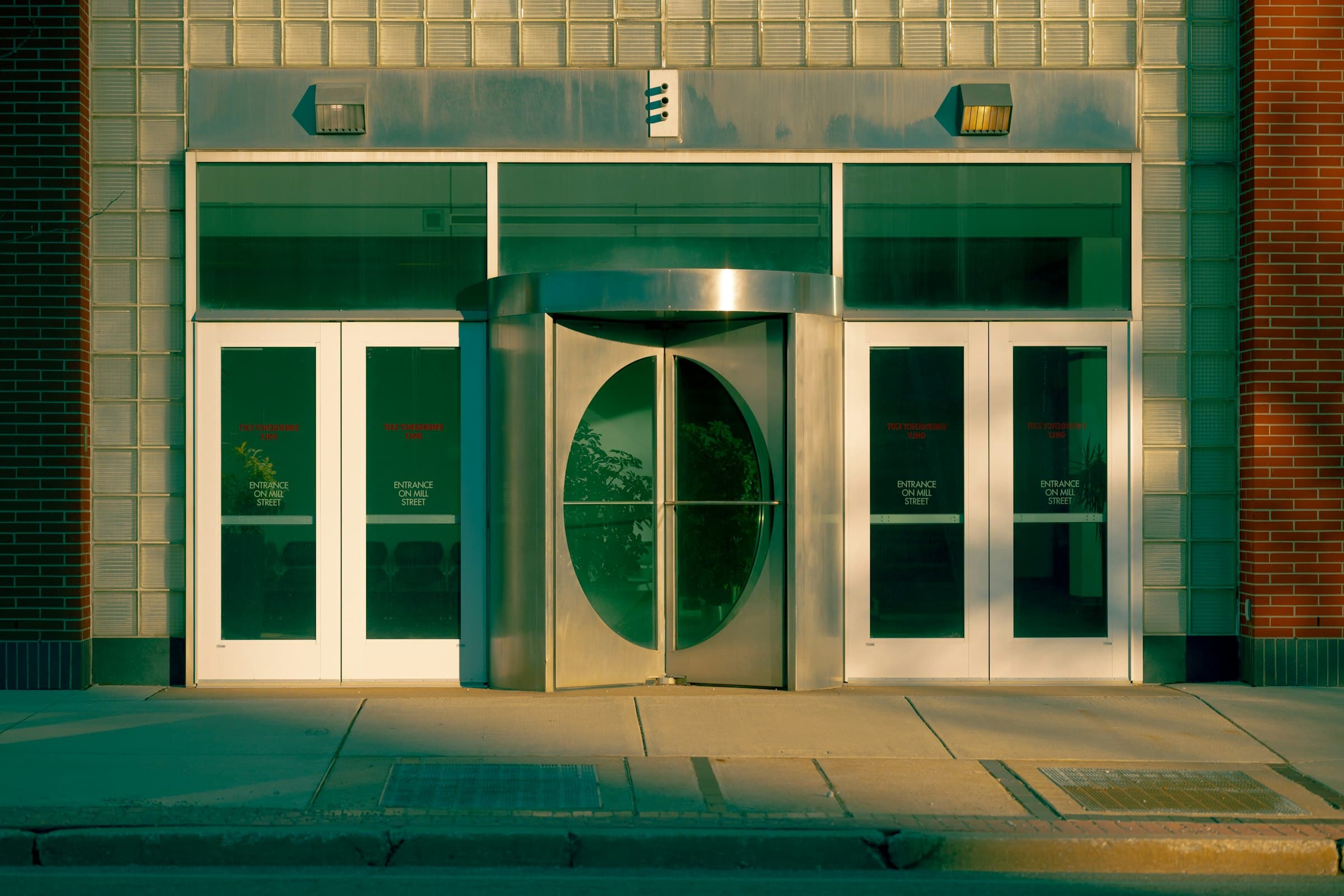 A revolving door between two pairs of glass doors at an office building entrance, lit gold and teal by low sun