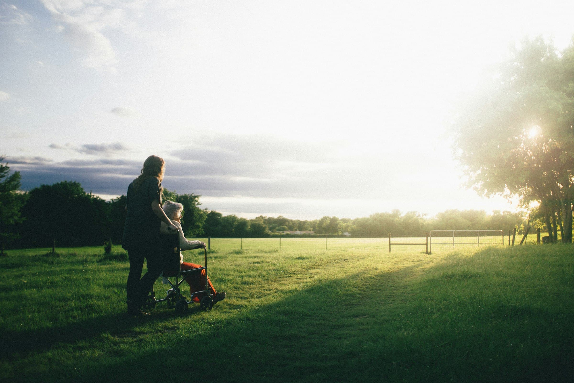 A caregiver pushing a person in a wheelchair across a field at golden hour