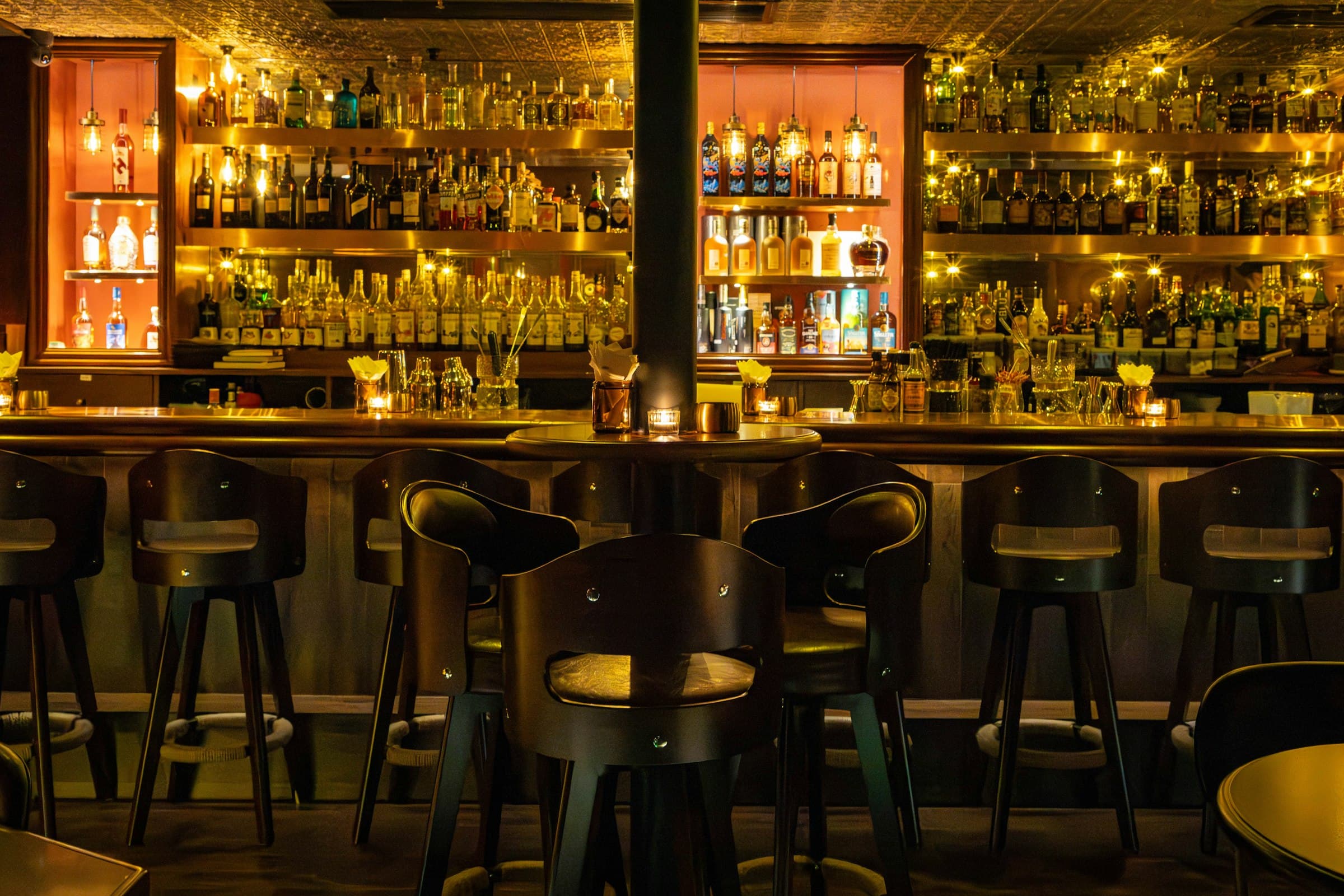 Bar stools in silhouette before shelves of backlit bottles glowing gold