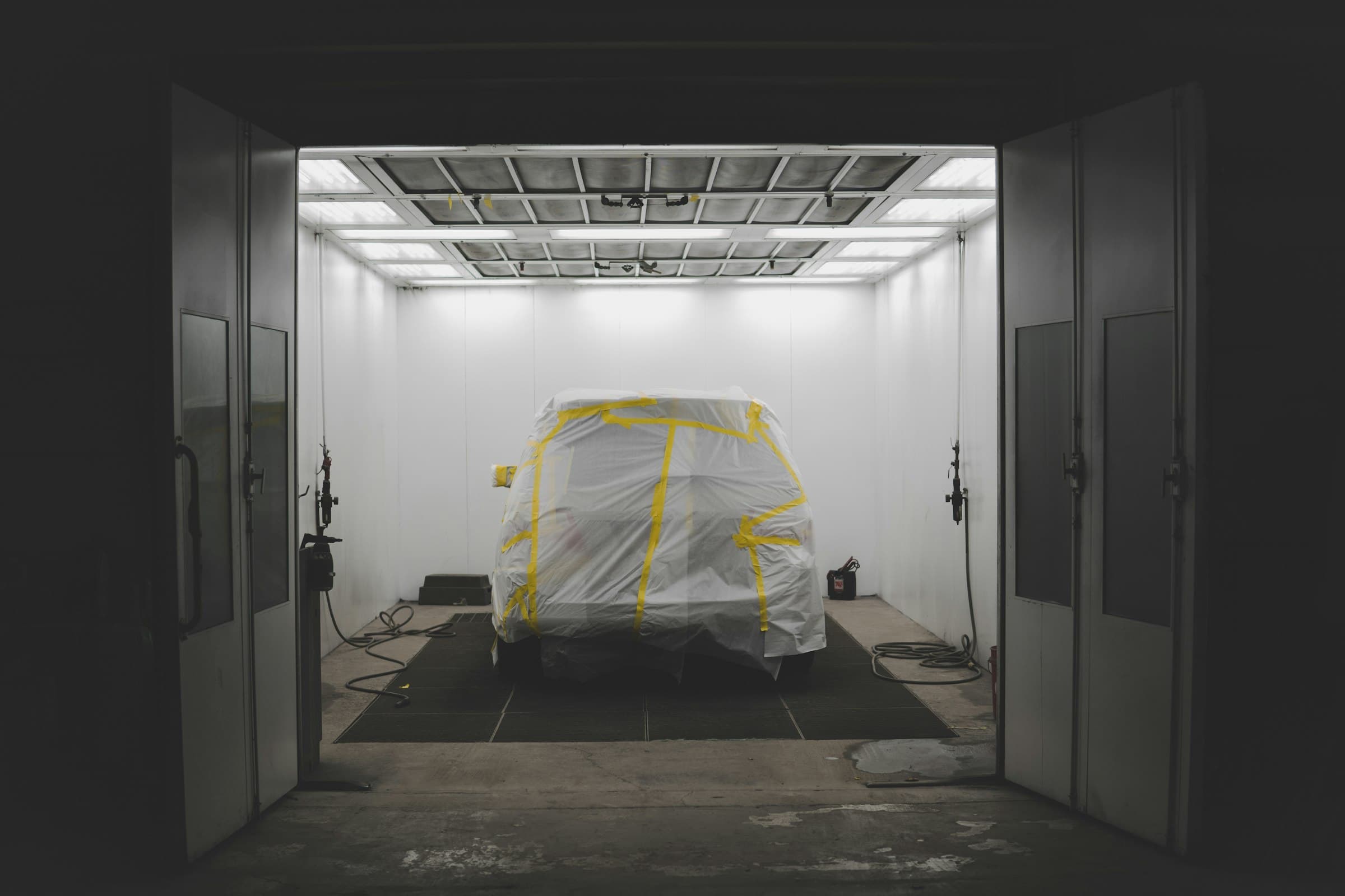 A car masked in plastic and tape inside a white-lit paint booth at a body shop