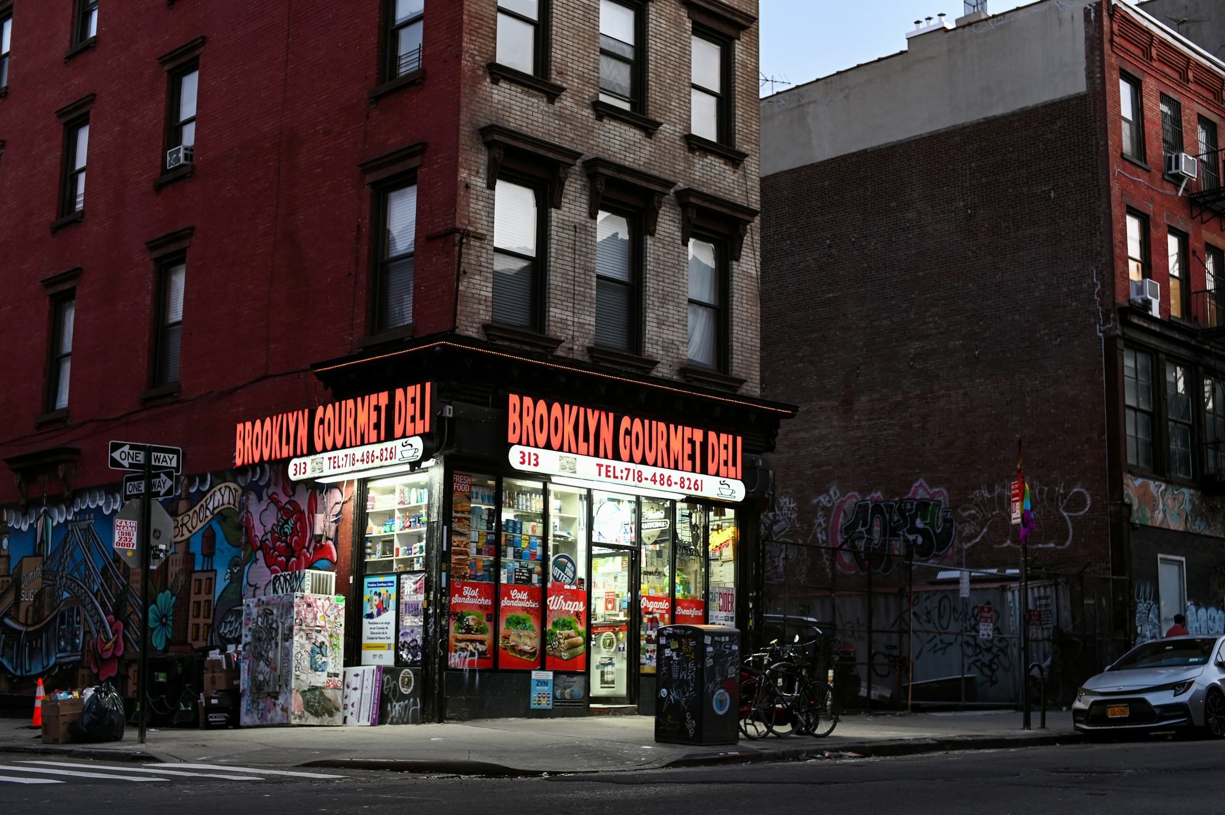 A corner deli with red signs on the ground floor of a brick building at dusk