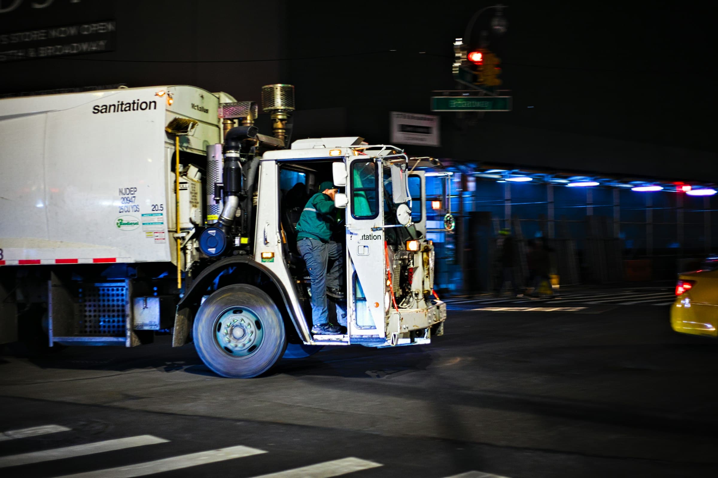 A sanitation truck at night, a worker climbing into the cab