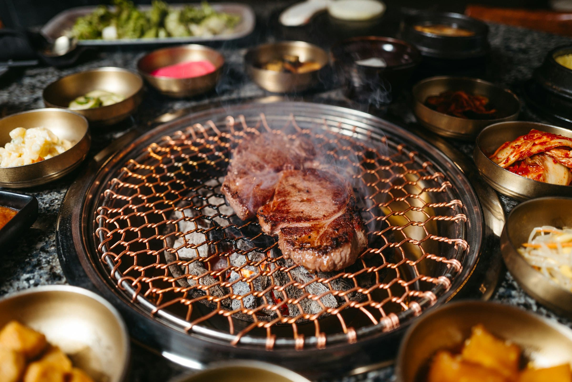 Meat on a tabletop barbecue grill surrounded by small bowls of side dishes