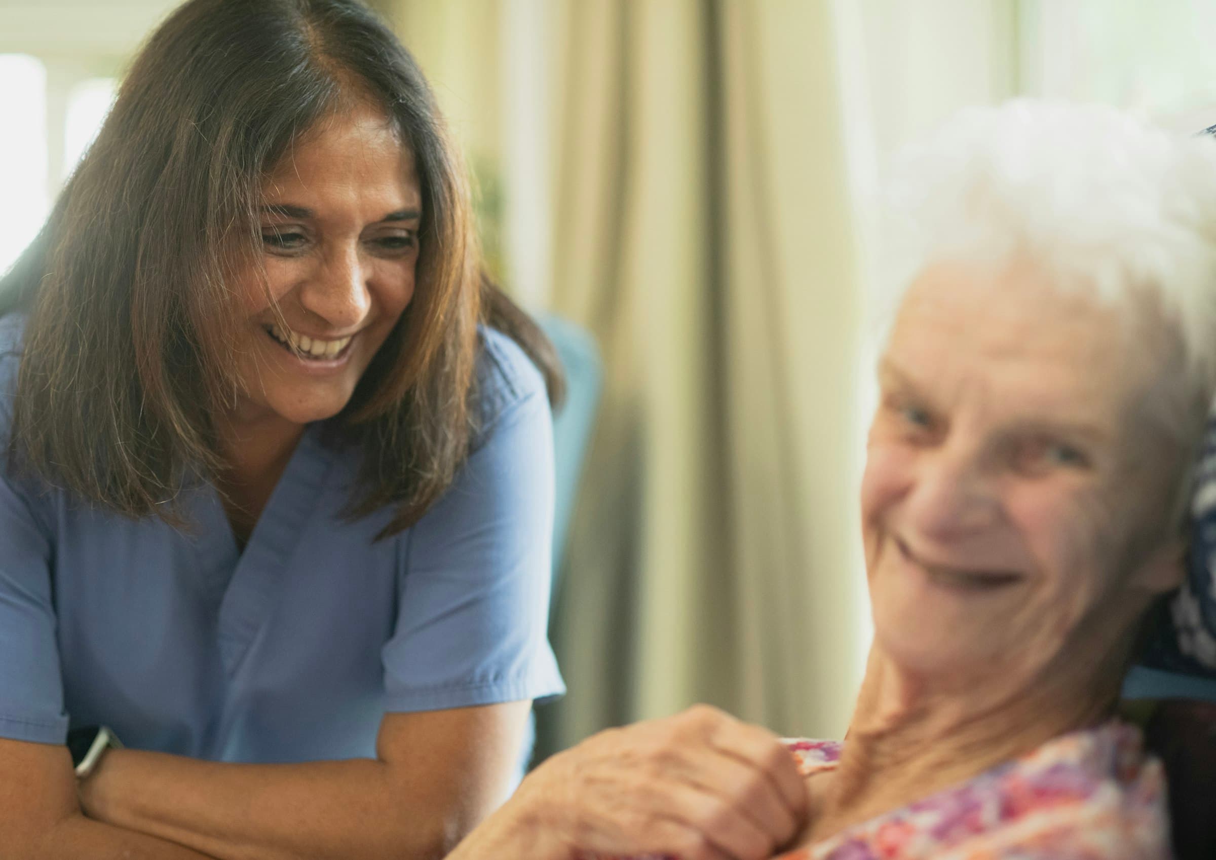 A nurse in blue scrubs smiling with an elderly resident in a sunlit room
