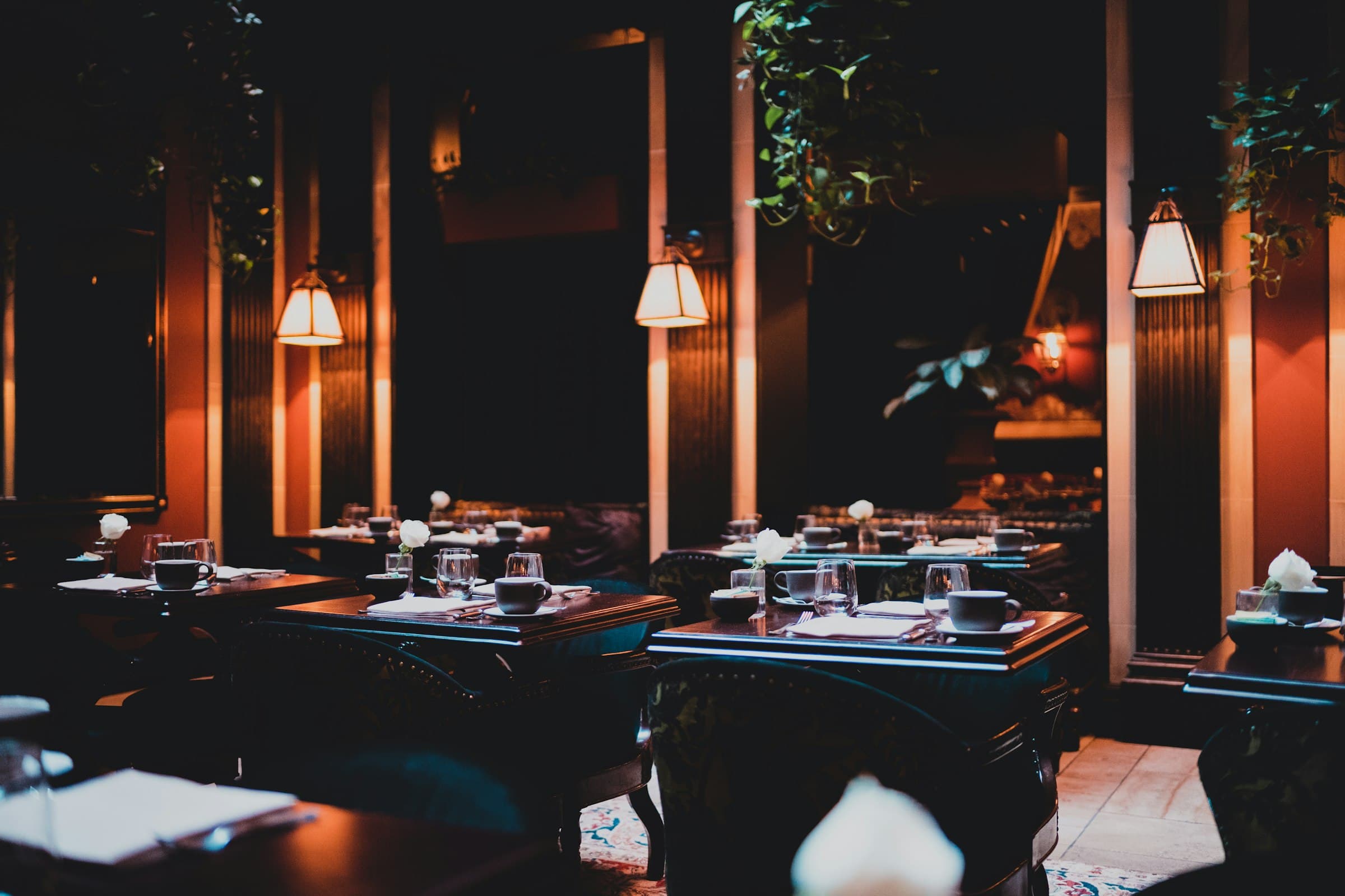 A restaurant dining room at night, set tables under warm wall lamps against dark teal and coral walls