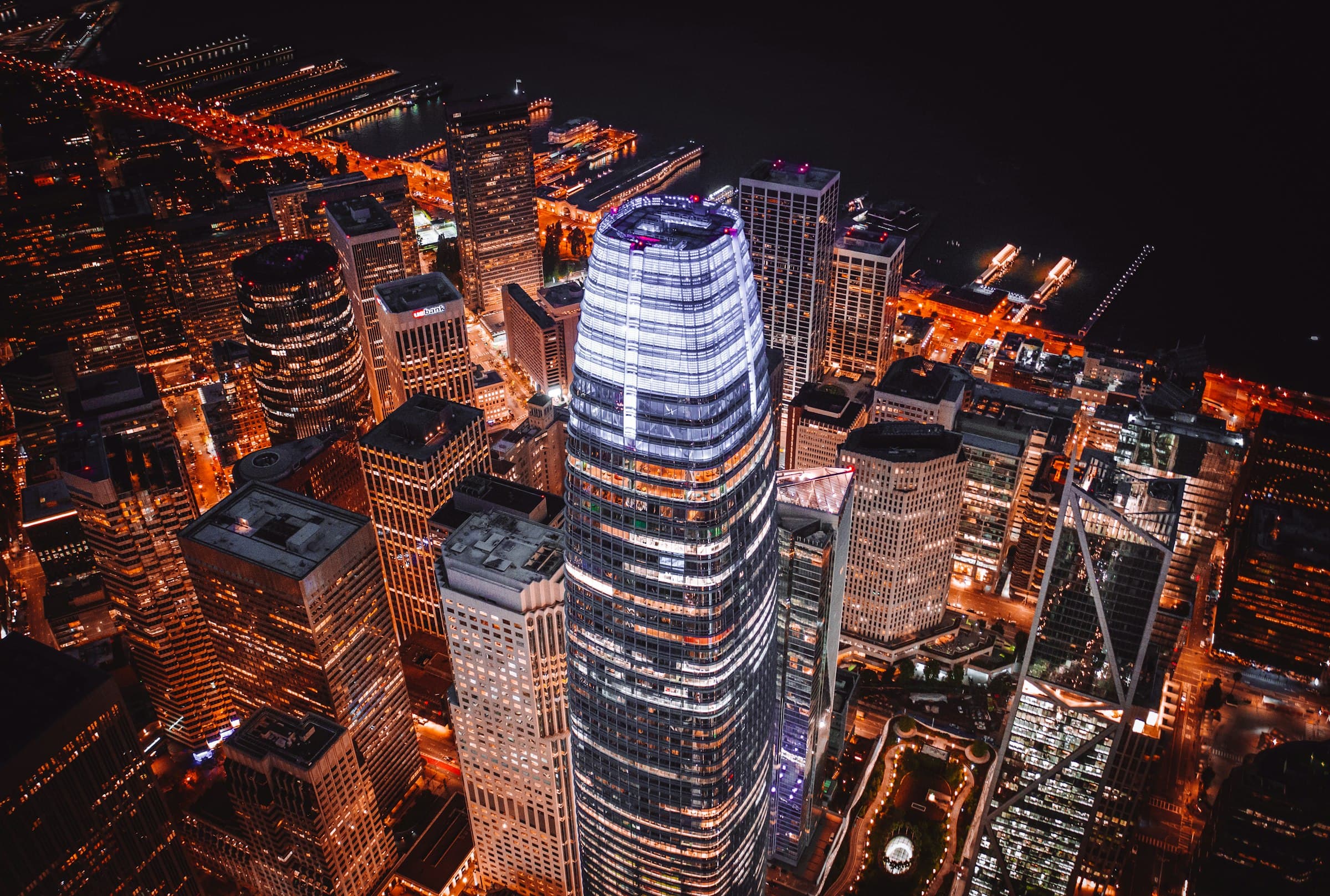 San Francisco's office towers from the air at night, the lit crown of the Salesforce Tower at the center and the Embarcadero piers beyond