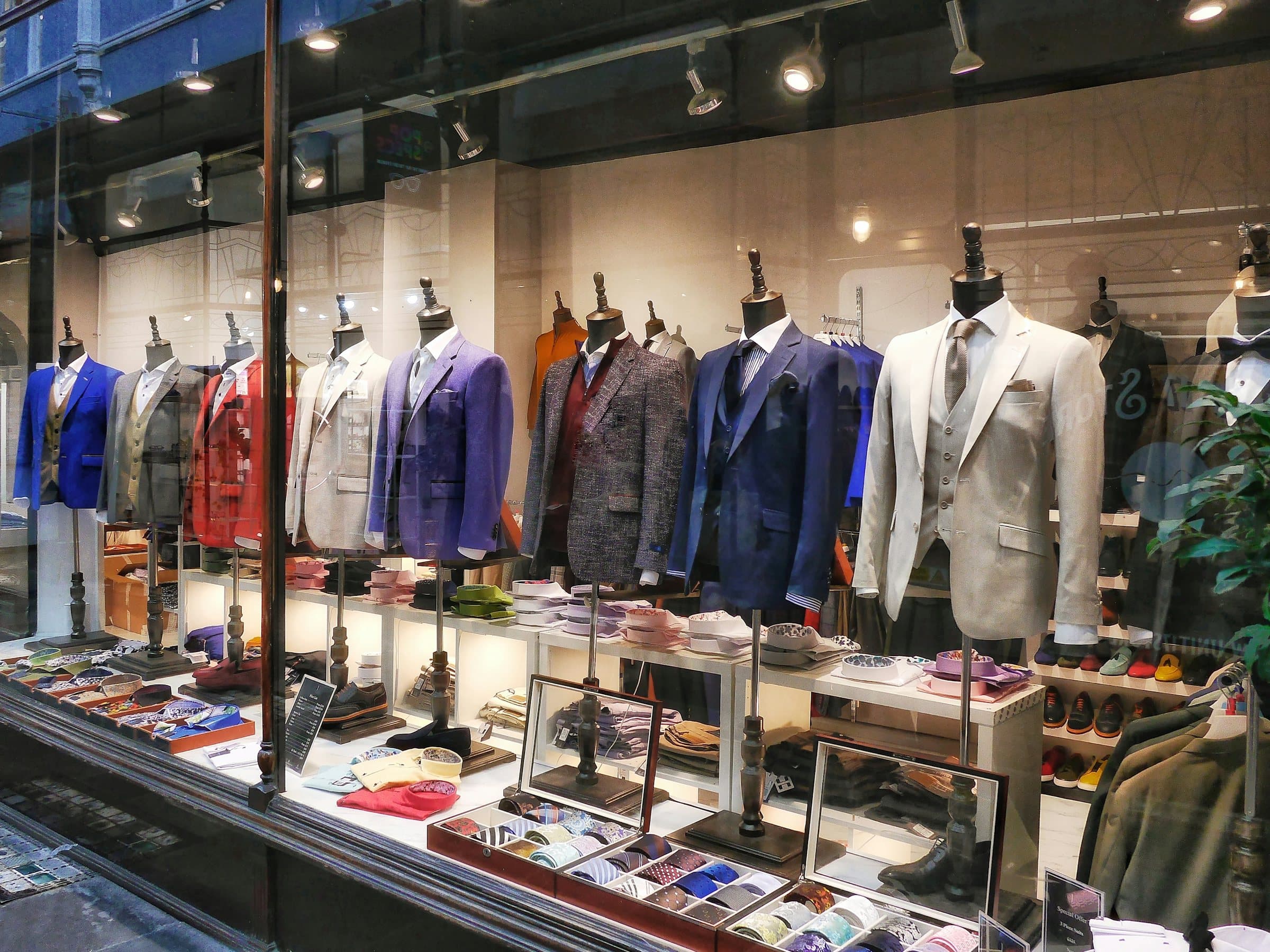 A menswear shop window with a row of jackets and suits on dress forms, boxes of ties and shoes on the shelves below